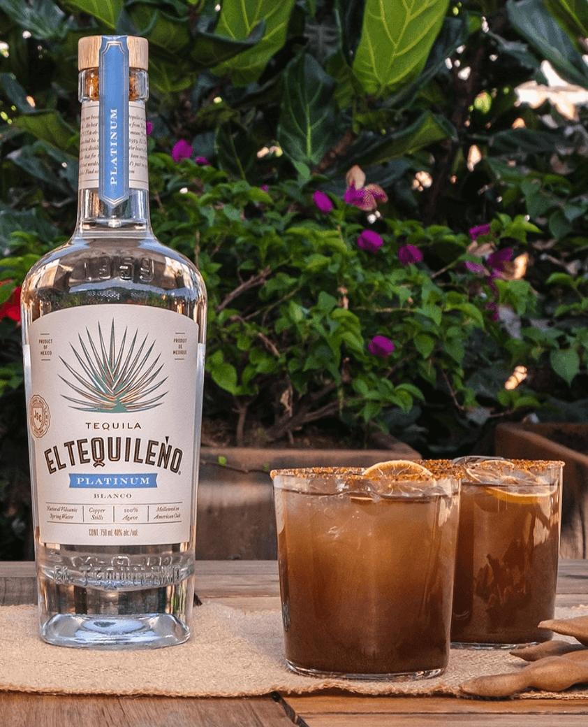 El Tequileño Platinum Blanco Tequila bottle with cocktails, surrounded by greenery and colorful flowers.