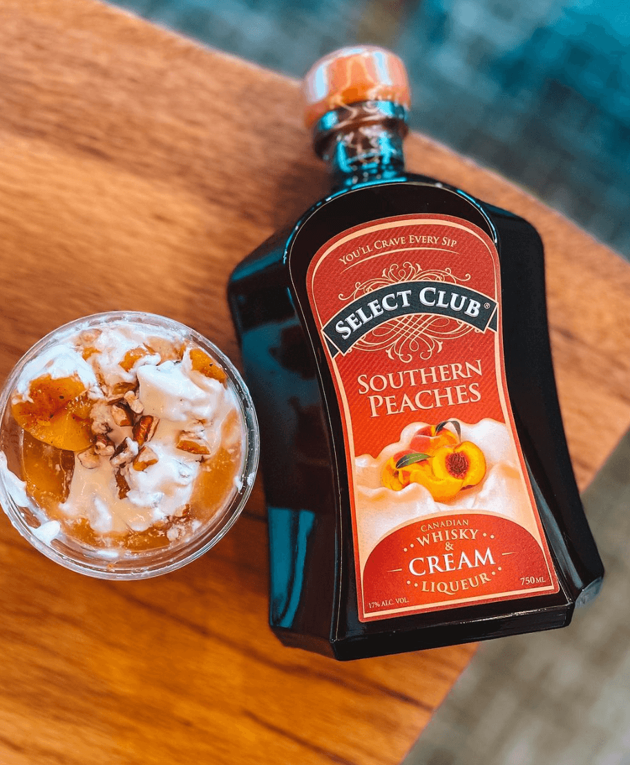 Select Club Southern Peaches whisky cream liqueur bottle beside a dessert glass topped with peaches and cream.