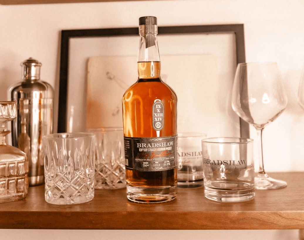 Bradshaw Kentucky Straight Bourbon whiskey bottle displayed on a wooden shelf with crystal glasses.