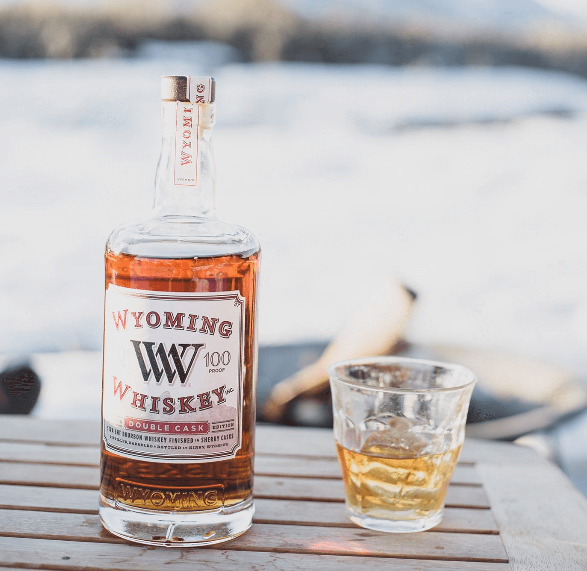 Bottle of Wyoming Whiskey with a glass of bourbon on a wooden table against a snowy background.