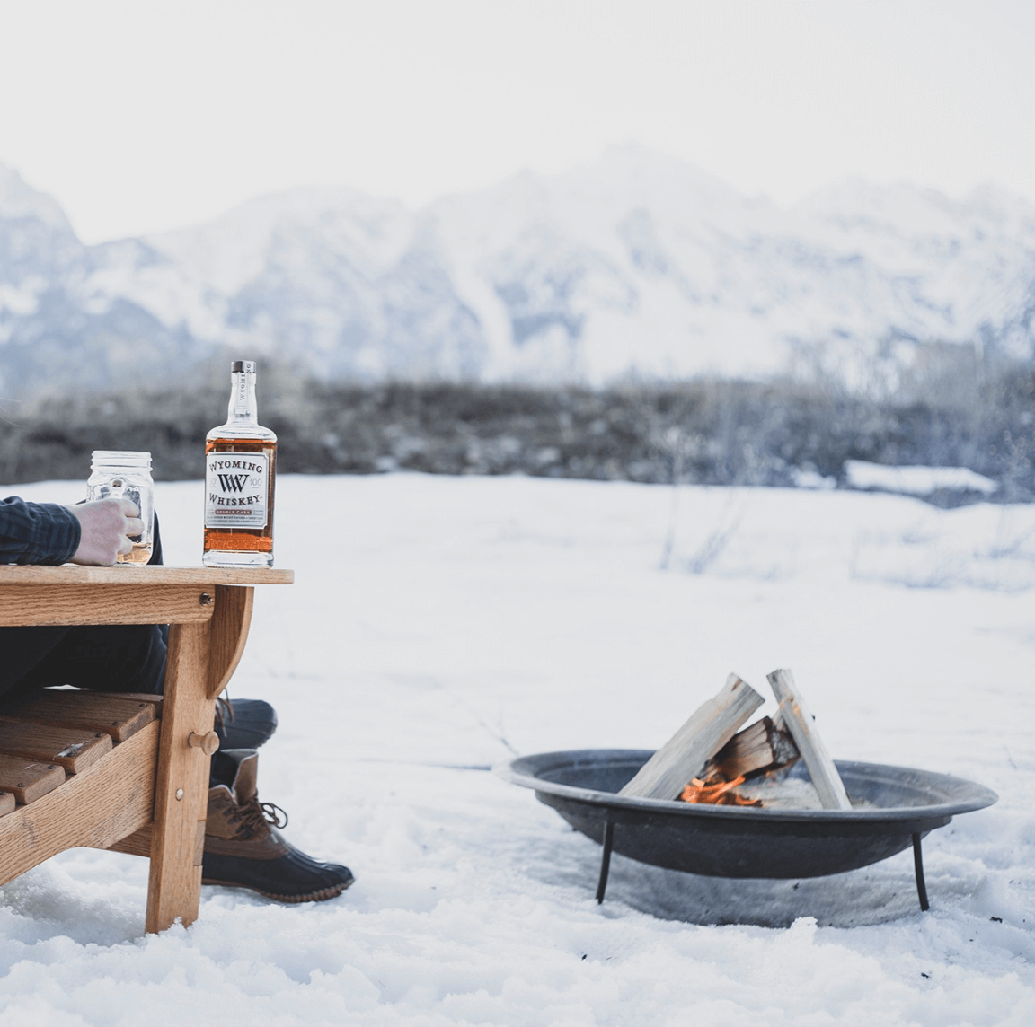 Person sitting by a fire in the snow, enjoying Wyoming Small Batch Bourbon Whiskey with mountains in the background.