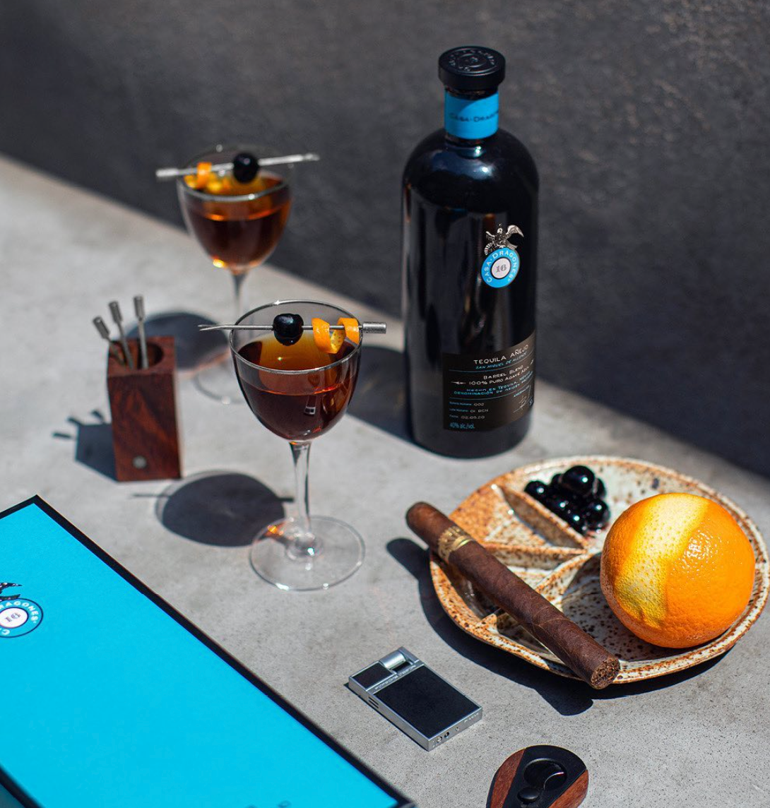 Casa Dragones Añejo Barrel Blend tequila bottle, cocktails, cigar, orange slice, and accessories on a stylish table.