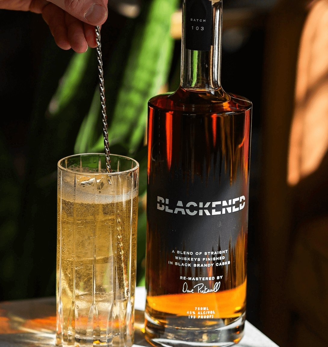 A bottle of Blackened American Whiskey with a glass, showcasing its rich color and branding, perfect for whiskey enthusiasts.