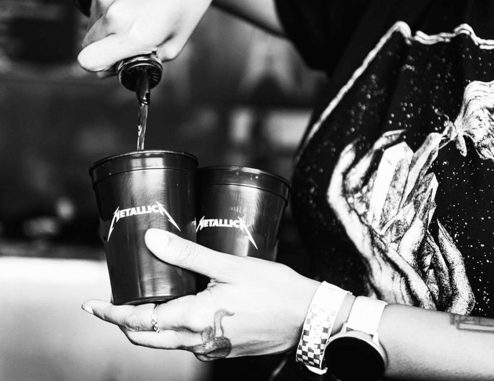 Person pouring Blackened American Whiskey into Metallica branded cups at a concert.