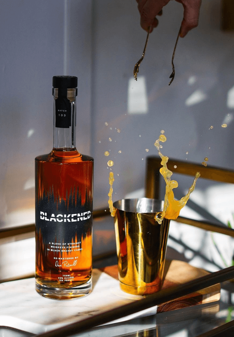 Bottle of Blackened American Whiskey beside a shaken cocktail in a gold shaker with splashes.