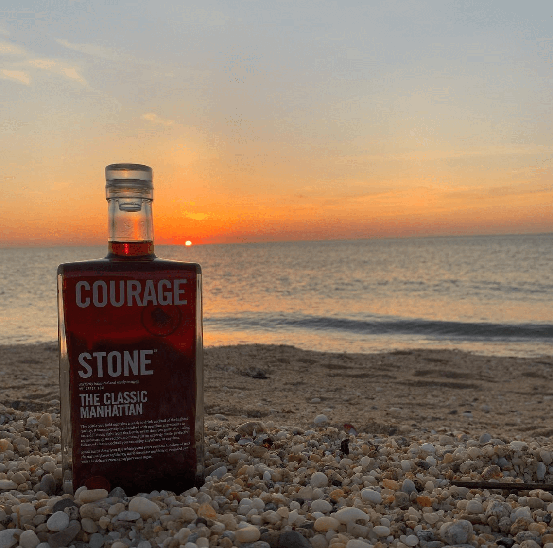 Courage + Stone Classic Manhattan cocktail bottle on a beach at sunset, highlighting its rich flavor and craft cocktail appeal.
