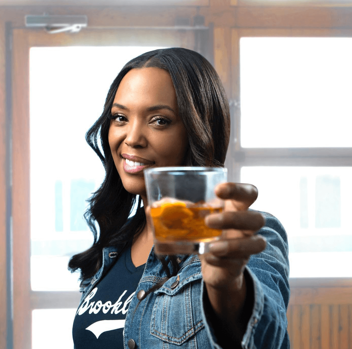 Aisha Tyler raising a glass of cocktail, showcasing Courage+Stone Classic Manhattan, smiling in a cozy setting.