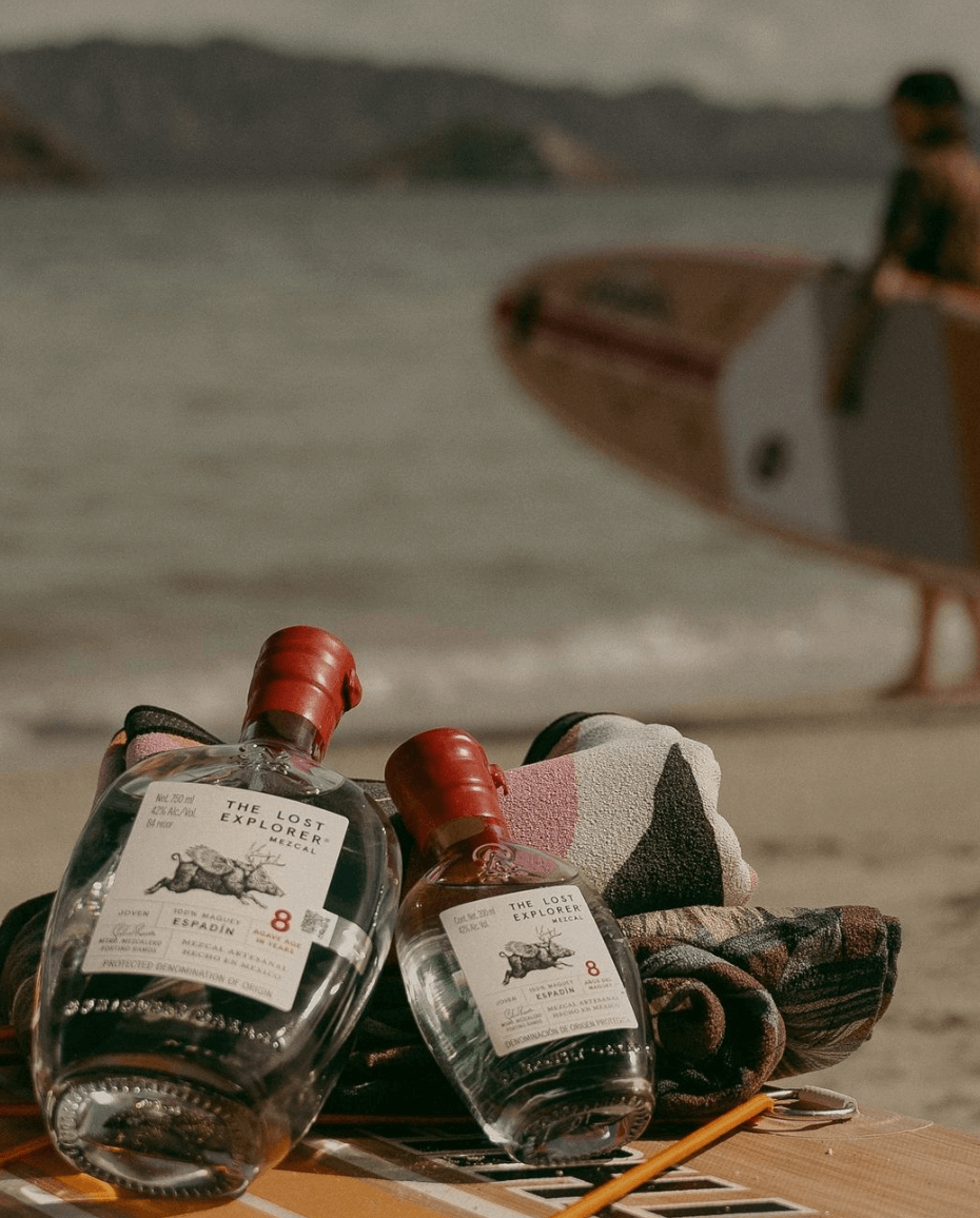The Lost Explorer Espadín 8 Year Mezcal bottles on the beach with surf equipment in the background.