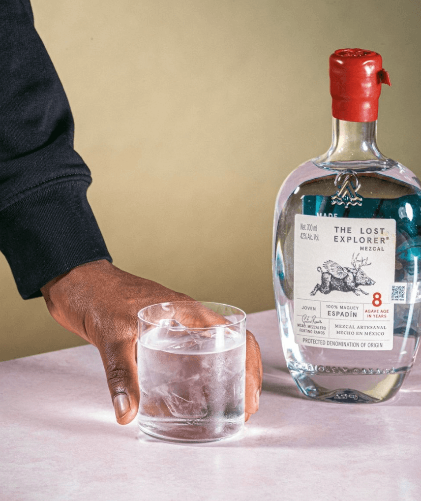 Hand reaching for a glass of ice next to The Lost Explorer Espadín 8 Year Mezcal bottle.