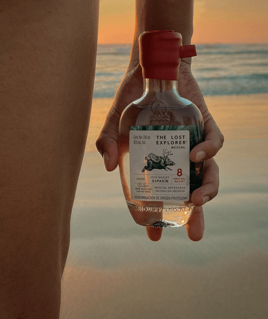 Hand holding The Lost Explorer Espadín 8 Year Mezcal bottle on the beach at sunset.
