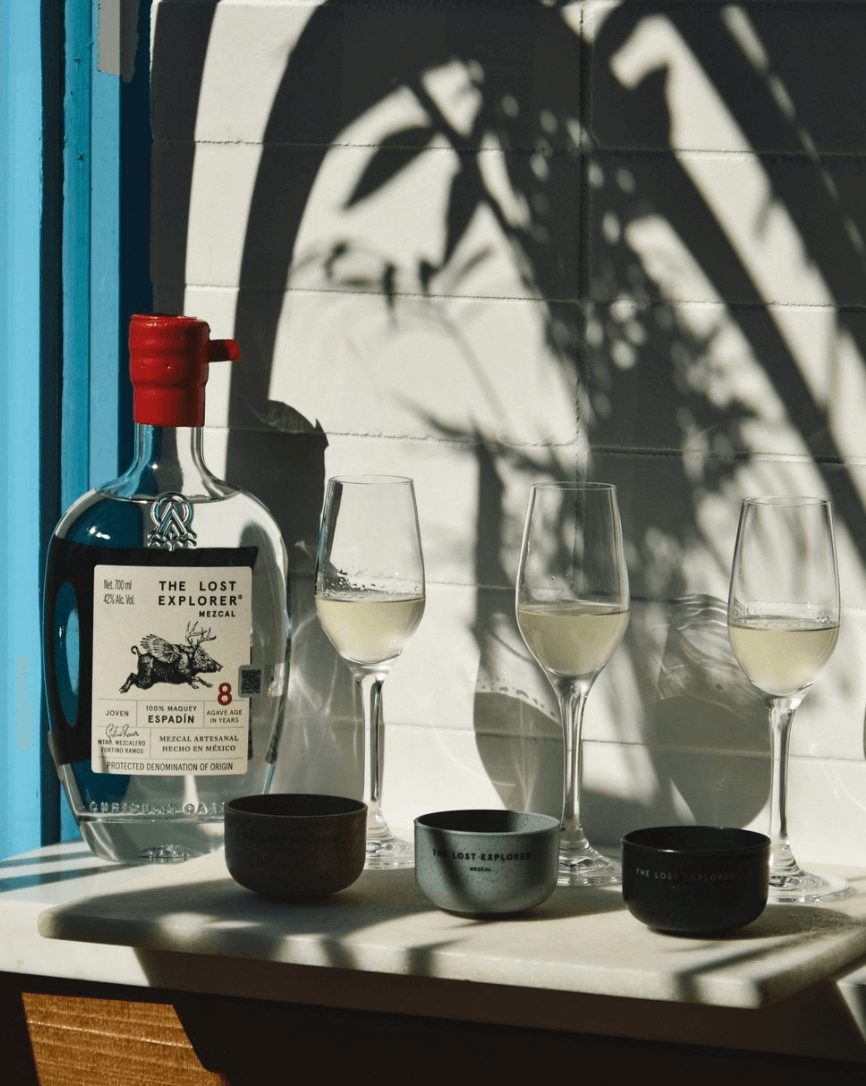 The Lost Explorer Espadín 8 Year Mezcal bottle with tasting glasses and shadows on a sunny table.