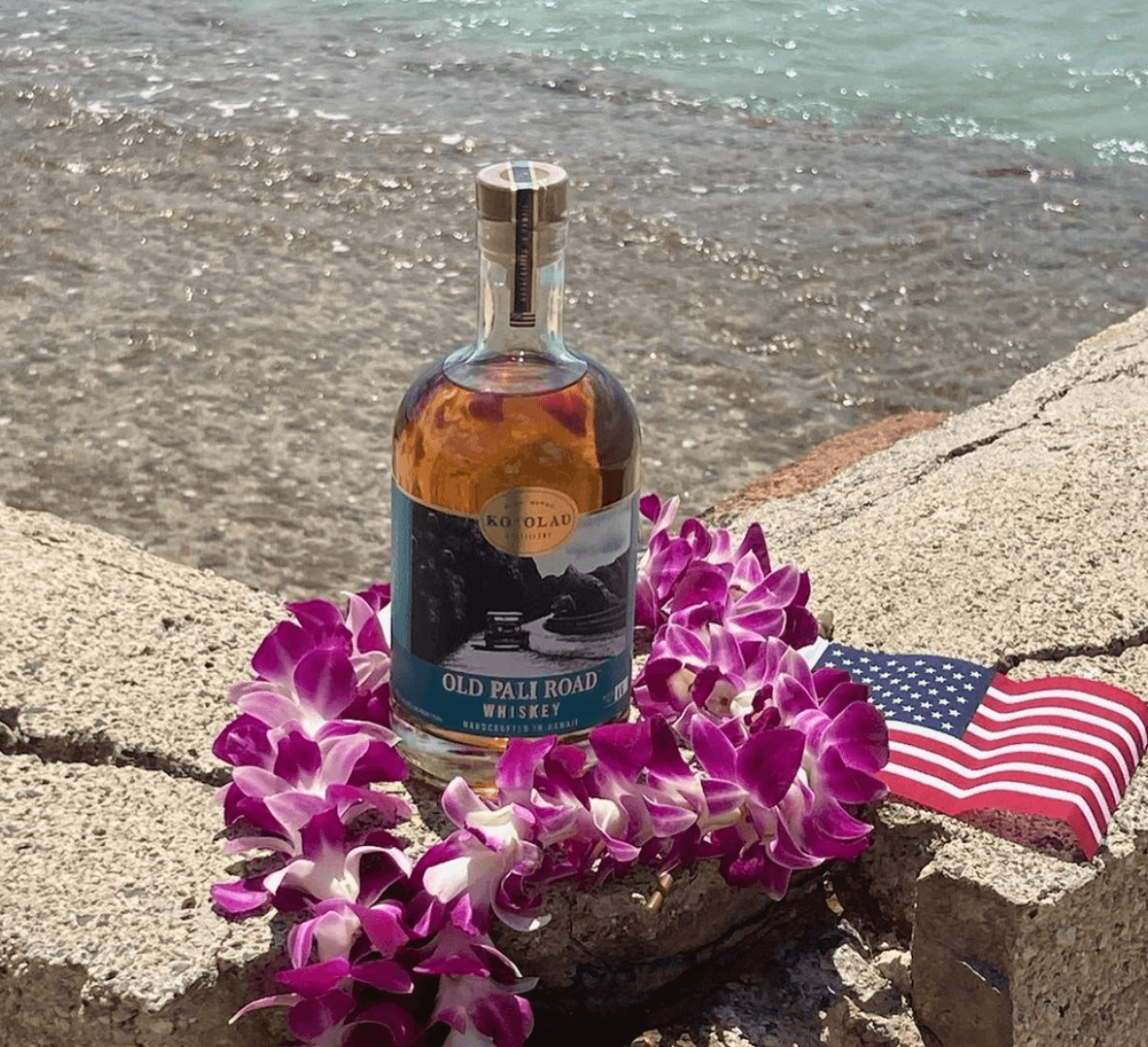 Old Pali Road Whiskey bottle surrounded by purple flowers and an American flag on a rocky beach.