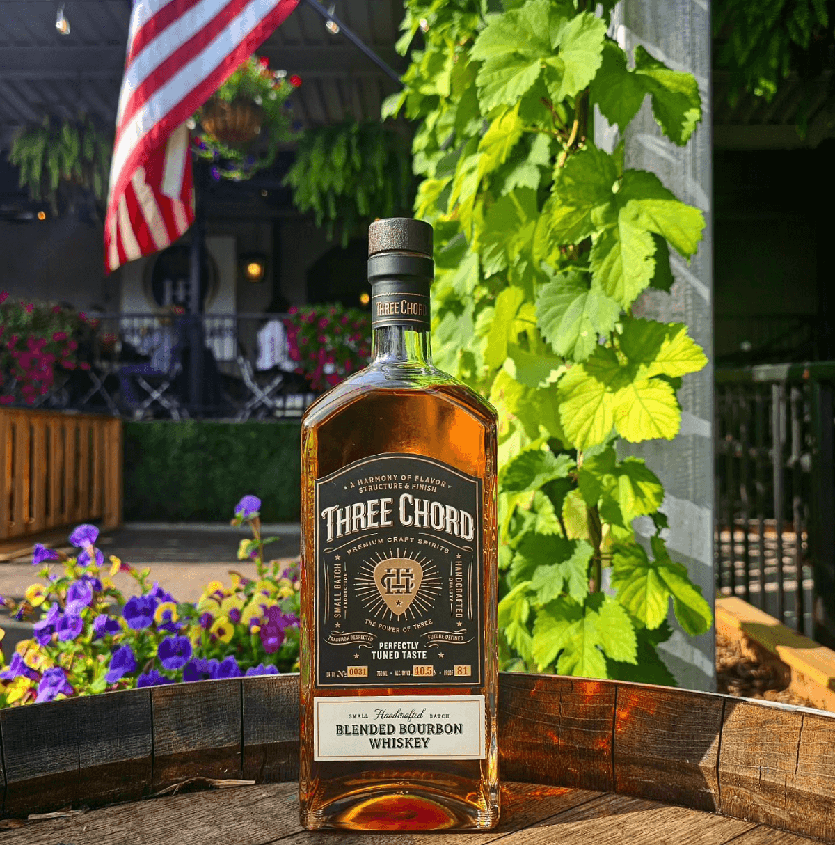 Three Chord Small Batch Blended Bourbon whiskey bottle in a garden setting with vibrant flowers and greenery.