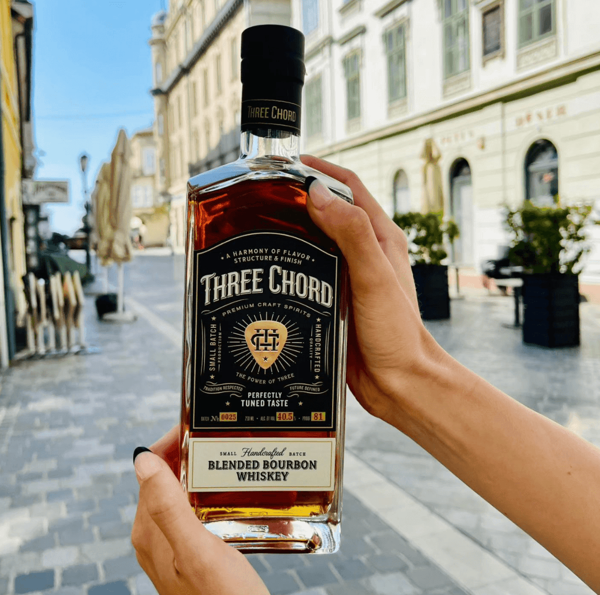 A person holding a bottle of Three Chord Small Batch Blended Bourbon in a charming street setting.