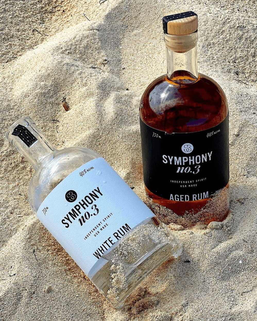 Symphony No.3 white rum and aged rum bottles nestled in sand, showcasing their sleek designs and labels.