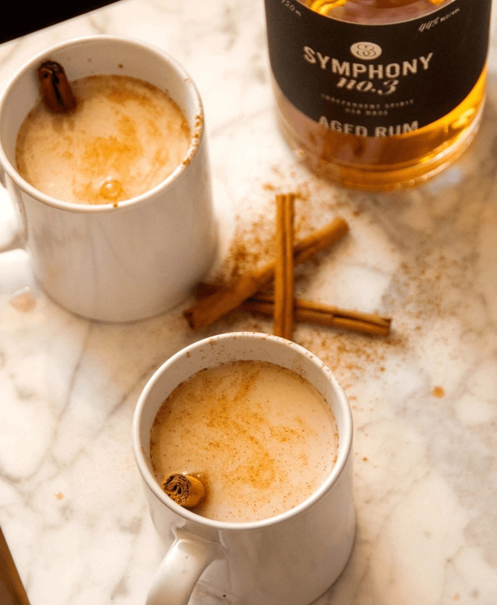 Two mugs of spiced drink with cinnamon sticks beside Symphony No.3 Aged Rum bottle on a marble surface.