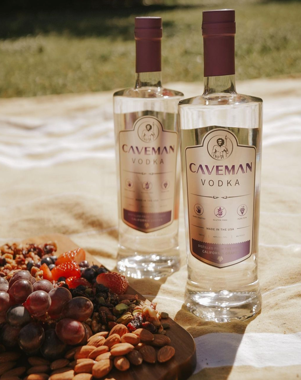 Caveman Vodka bottles on a picnic blanket with nuts and fruits, showcasing handcrafted, Paleo-friendly ingredients.