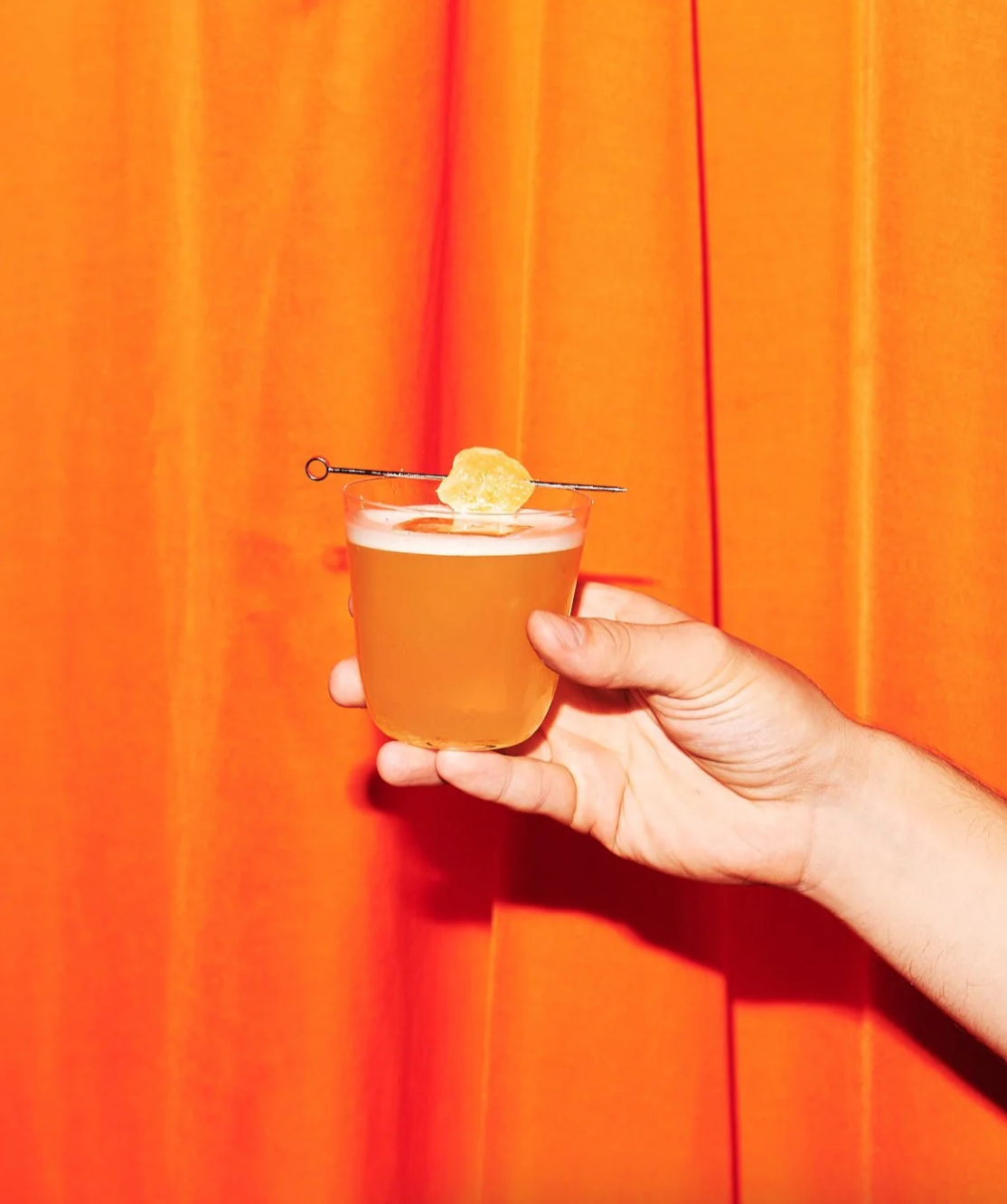 A hand holding a cocktail garnished with a slice of citrus, set against an orange curtain backdrop.