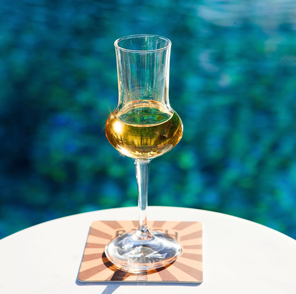 Glass of Batch 22 New American Aquavit Classic Gold resting on a coaster by a shimmering blue pool.