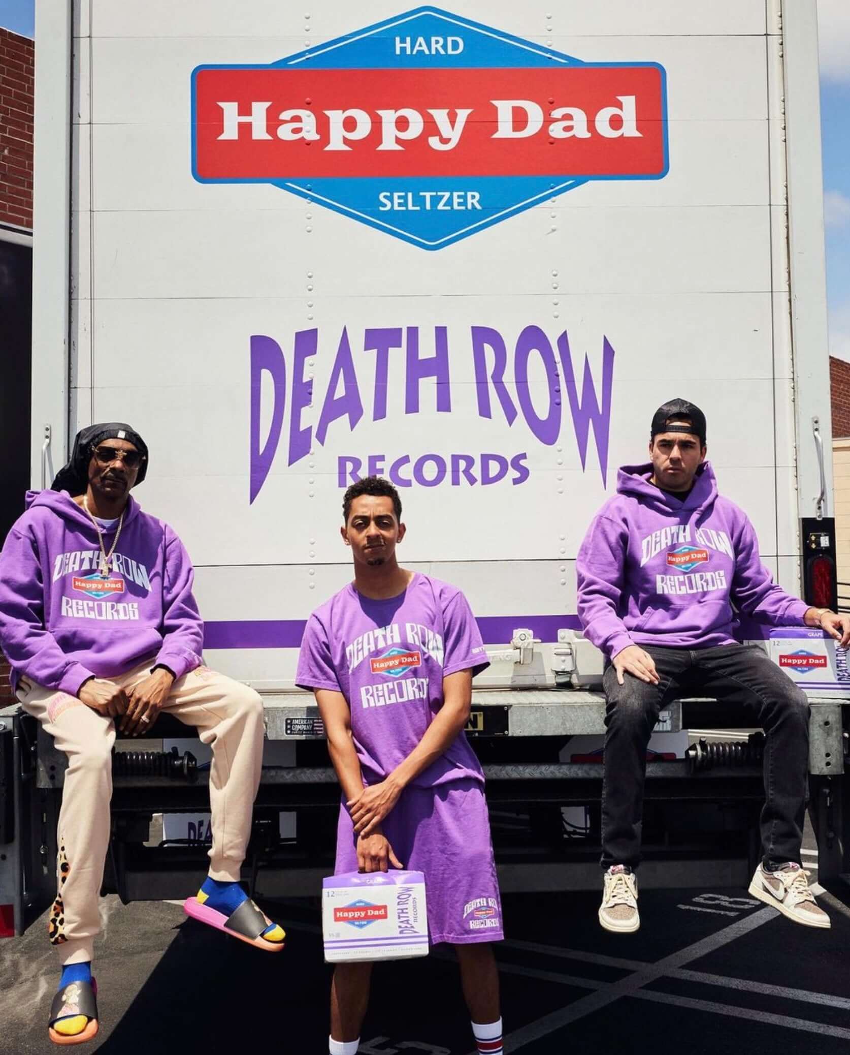 Three individuals in purple attire promoting Happy Dad x Death Row Records hard seltzer next to a branded truck.
