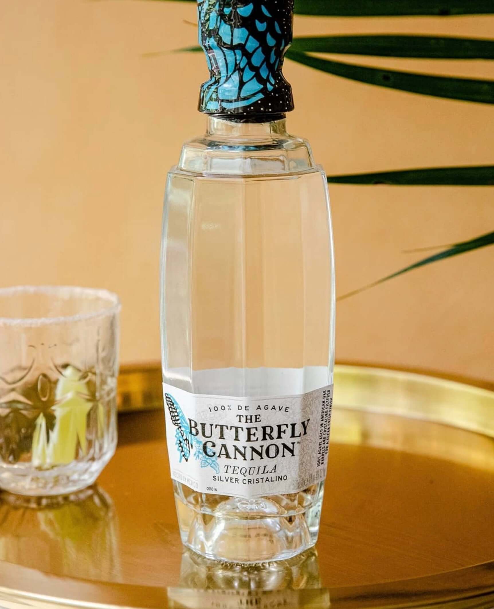 Butterfly Cannon Silver Cristalino tequila bottle with blue butterfly design, displayed on a gold tray.