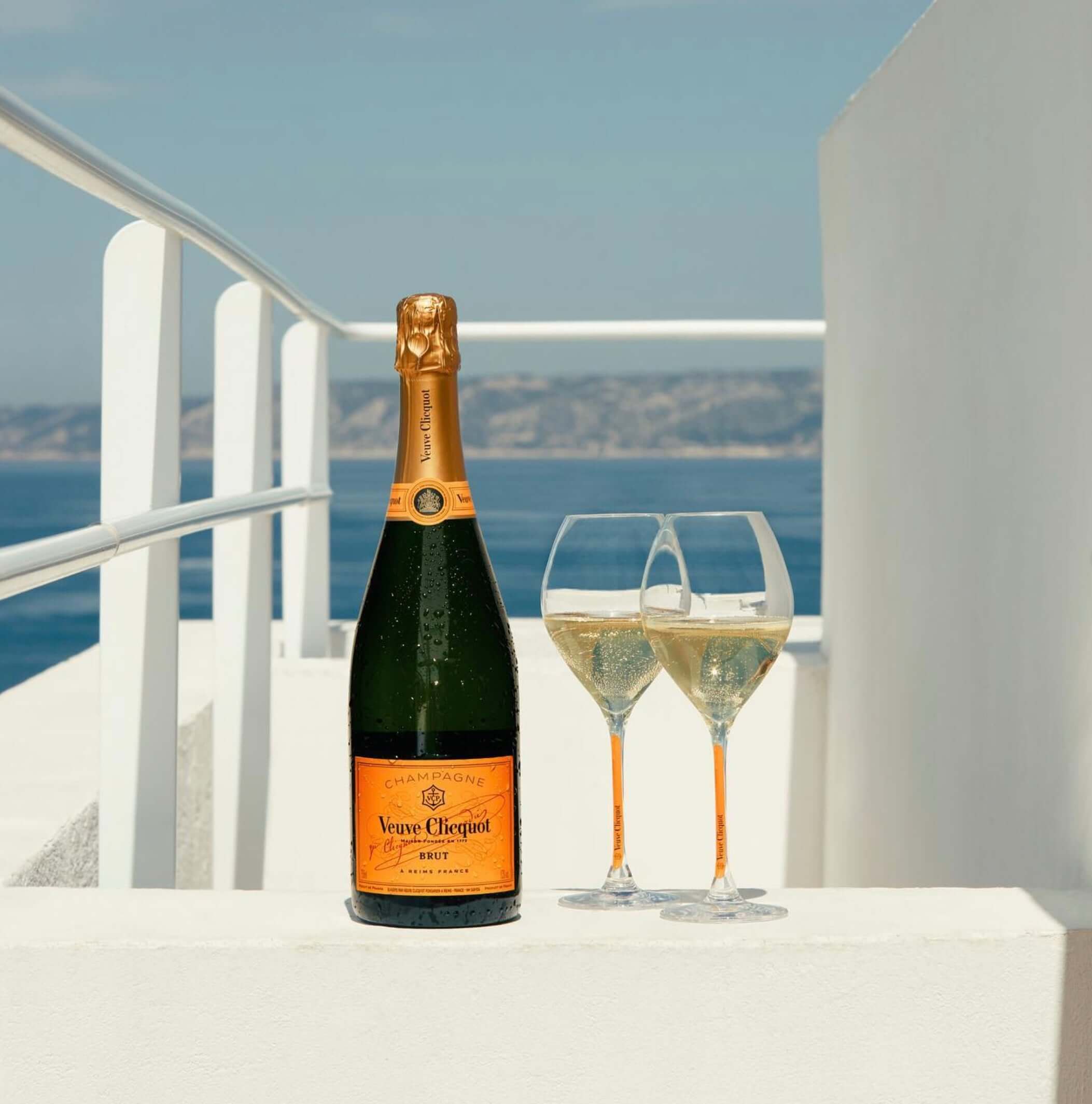Veuve Clicquot Brut Champagne bottle with two glasses on a sunlit terrace overlooking the ocean.