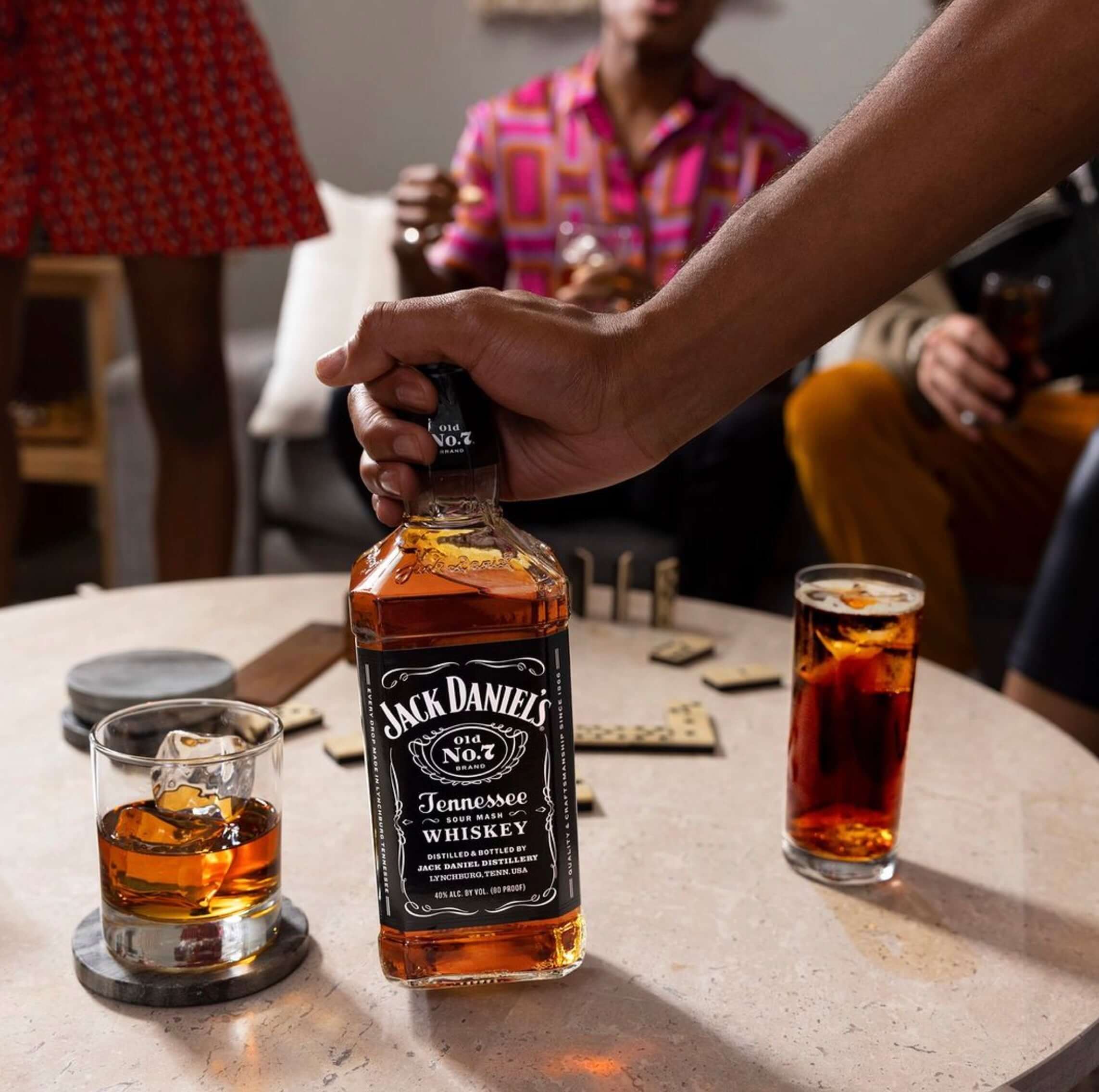 Hand holding a bottle of Jack Daniel's Tennessee Whiskey, with drinks and friends in a casual setting.