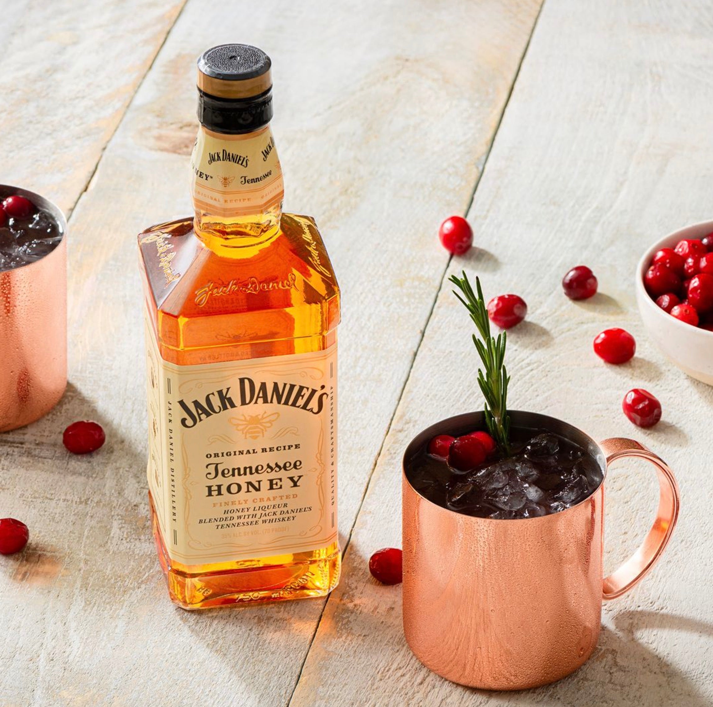 Jack Daniel's Tennessee Honey whiskey bottle next to a copper mug with drink and cranberries on a wooden surface.