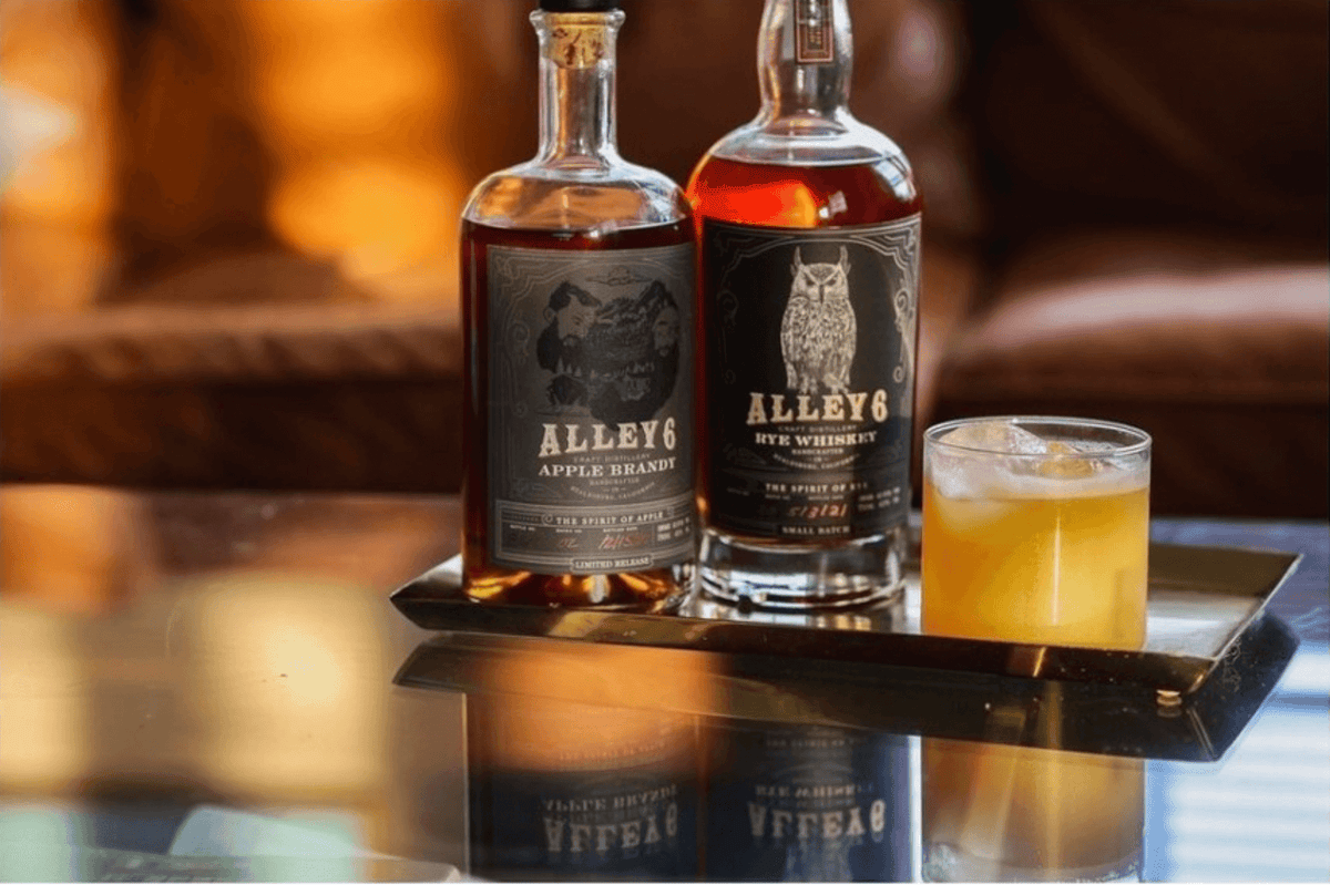 Two bottles of Alley 6 Apple Brandy and Rye Whiskey on a table with a glass of cocktail, showcasing rich amber colors.