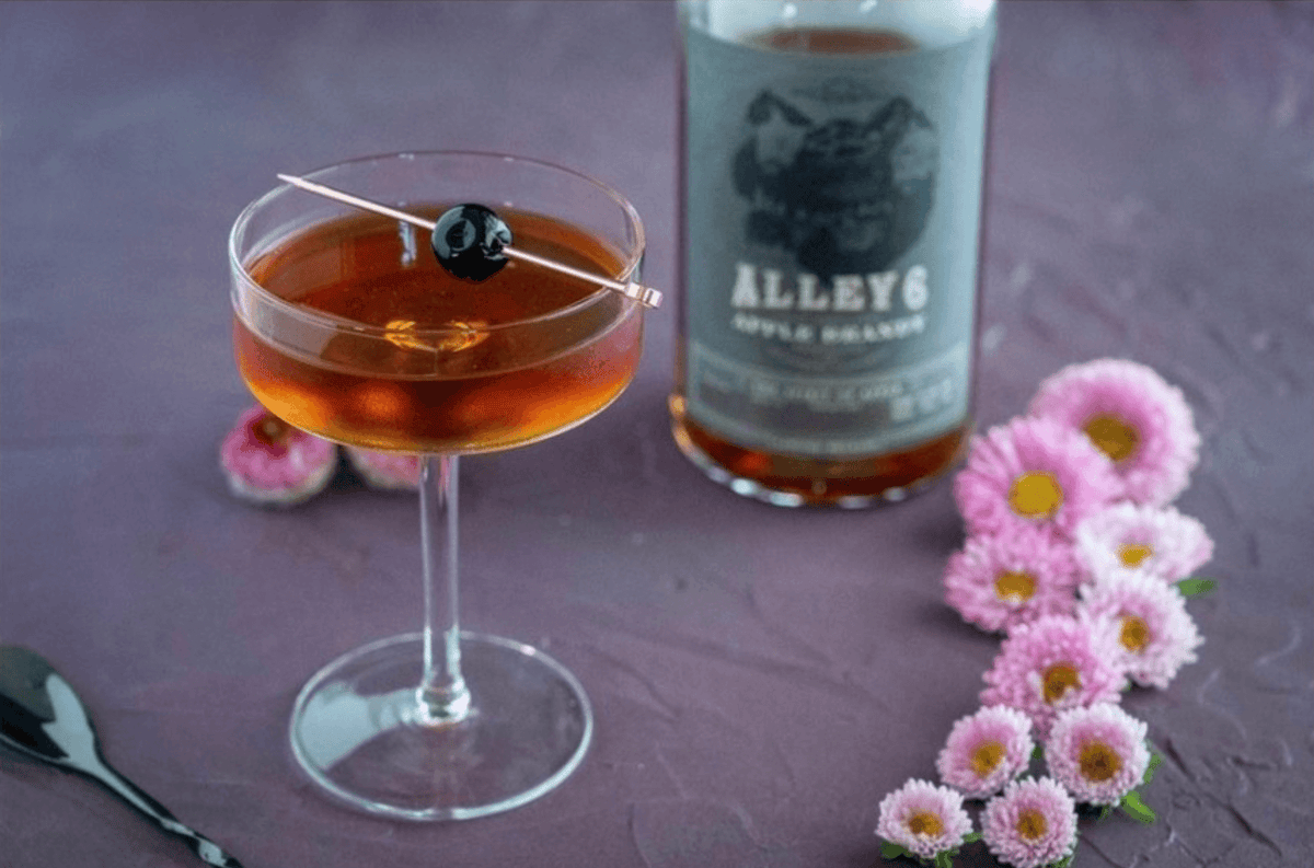 A cocktail with Alley 6 Apple Brandy, garnished with a cherry and surrounded by pink flowers.