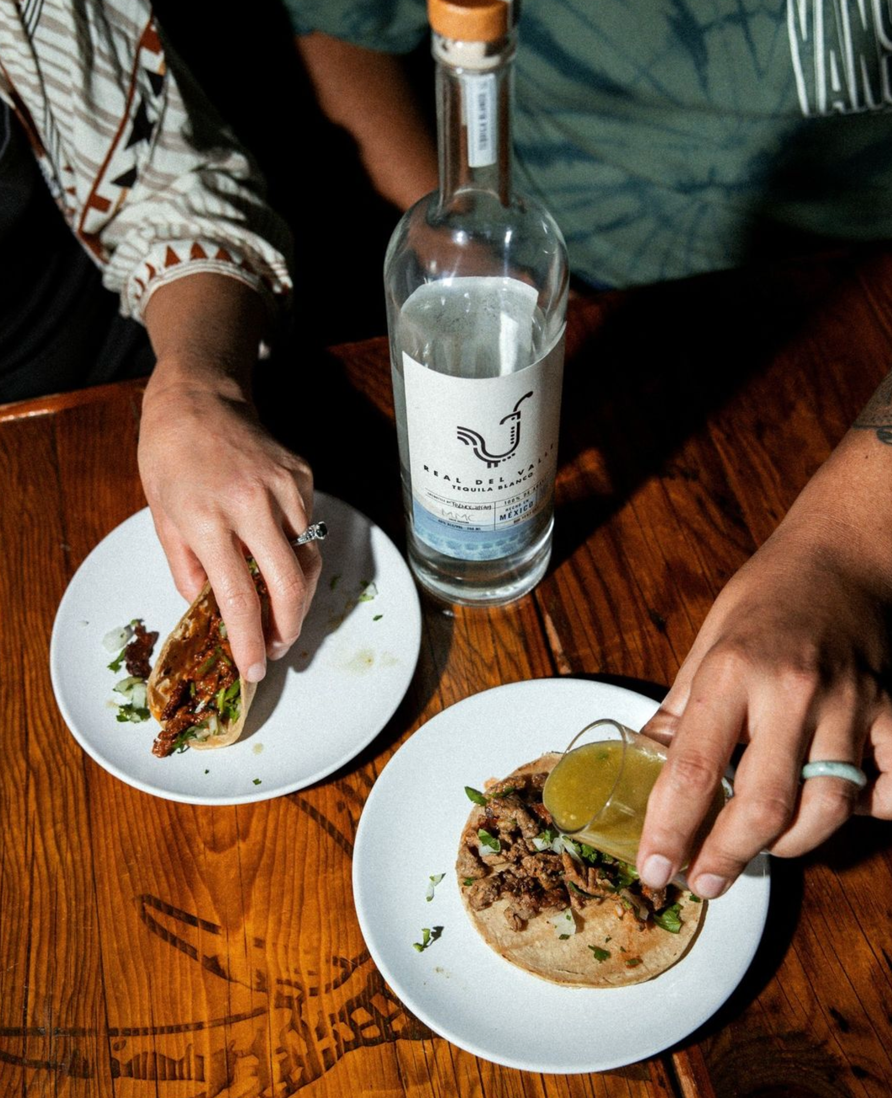 Two hands enjoying tacos with a bottle of Real Del Valle Blanco Tequila on a wooden table.