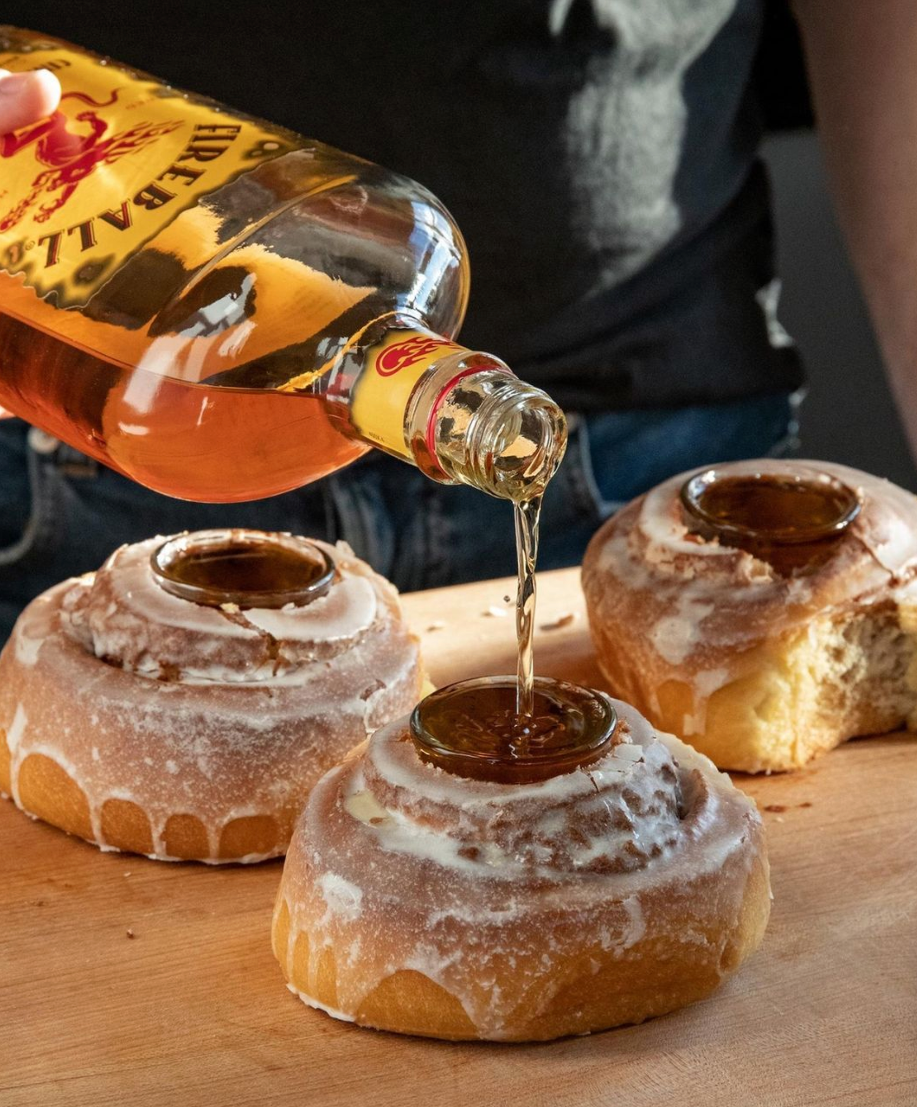 Pouring Fireball Cinnamon Whisky into cinnamon roll shots on a wooden surface.