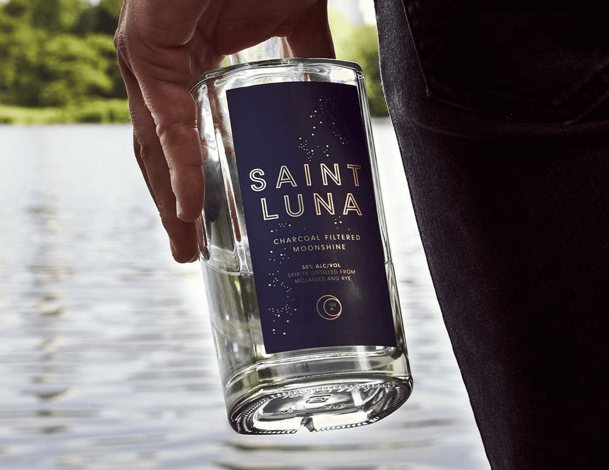 Saint Luna Charcoal Filtered Moonshine held by a person near a tranquil water backdrop, showcasing its elevated craft quality.