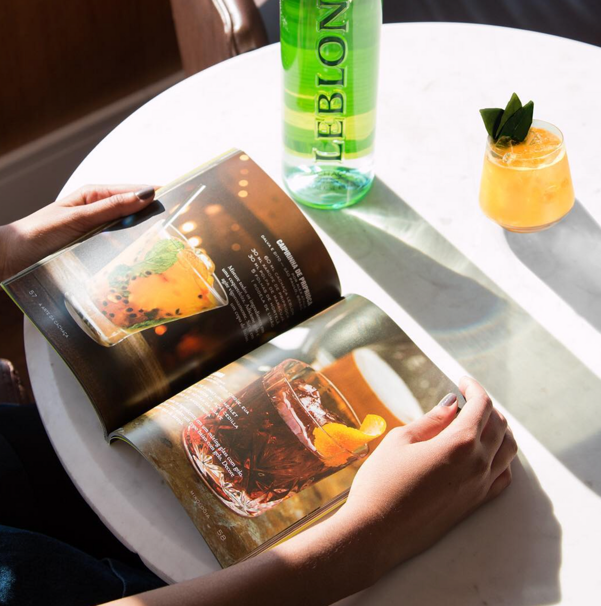 Person holding a cocktail recipe book next to a bottle of Leblon Cachaça and a refreshing cocktail.