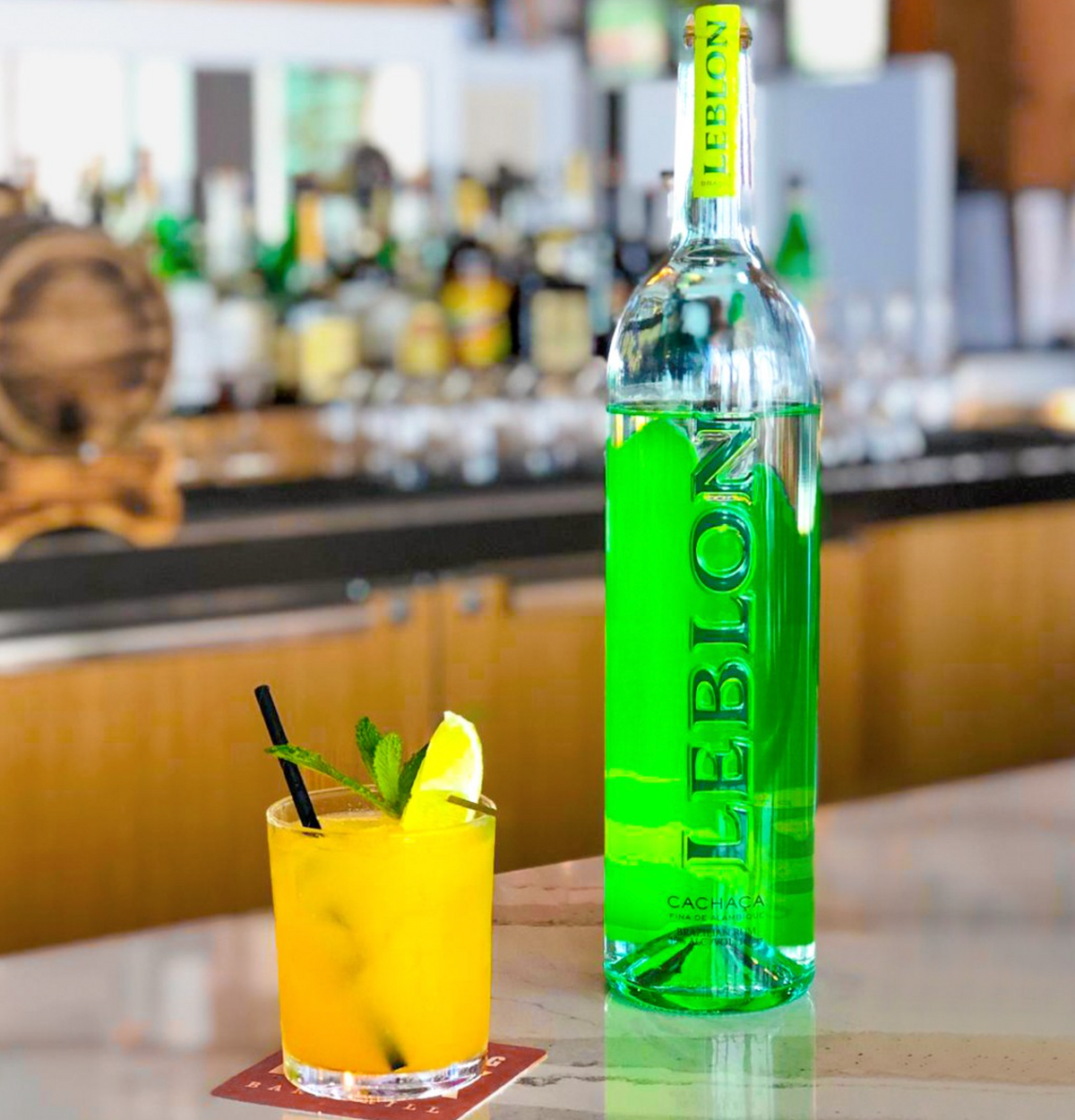 Bottle of Leblon Cachaça beside a vibrant cocktail with orange juice and lime, in a bar setting.