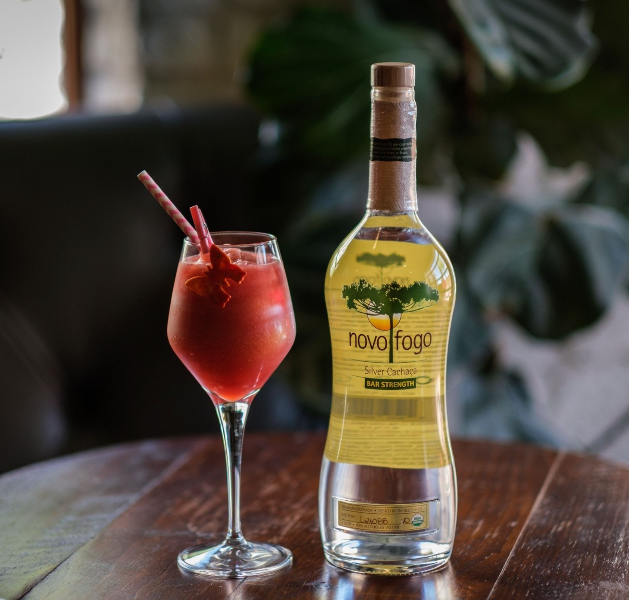 Novo Fogo Bar Strength Silver Cachaça 1L bottle next to a vibrant frozen cocktail on a wooden table.