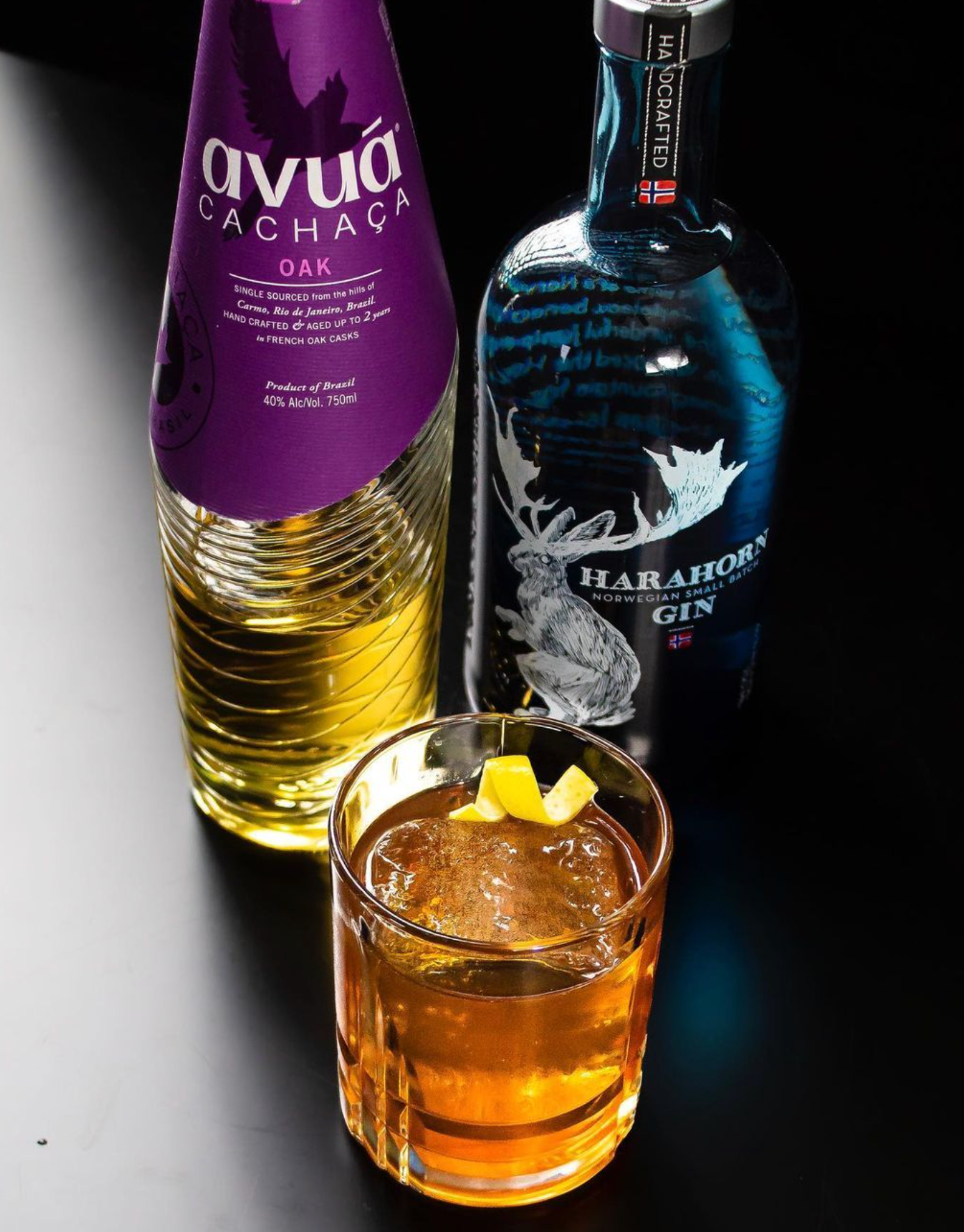 Avuá Oak Cachaça and Harahorn Gin displayed with a cocktail glass on a dark surface, highlighting premium spirits.