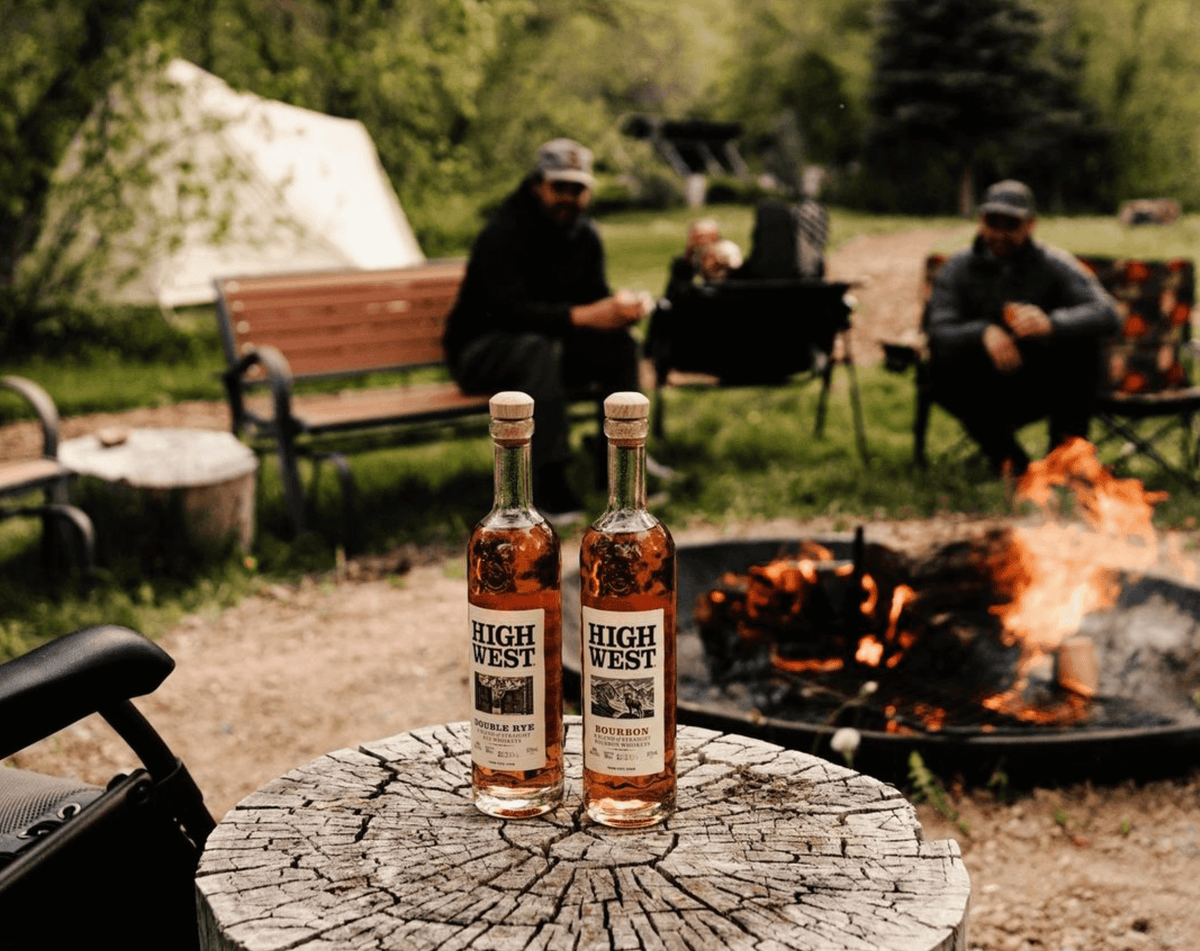 Two bottles of High West Bourbon on a wooden stump by a campfire, surrounded by nature and friends.