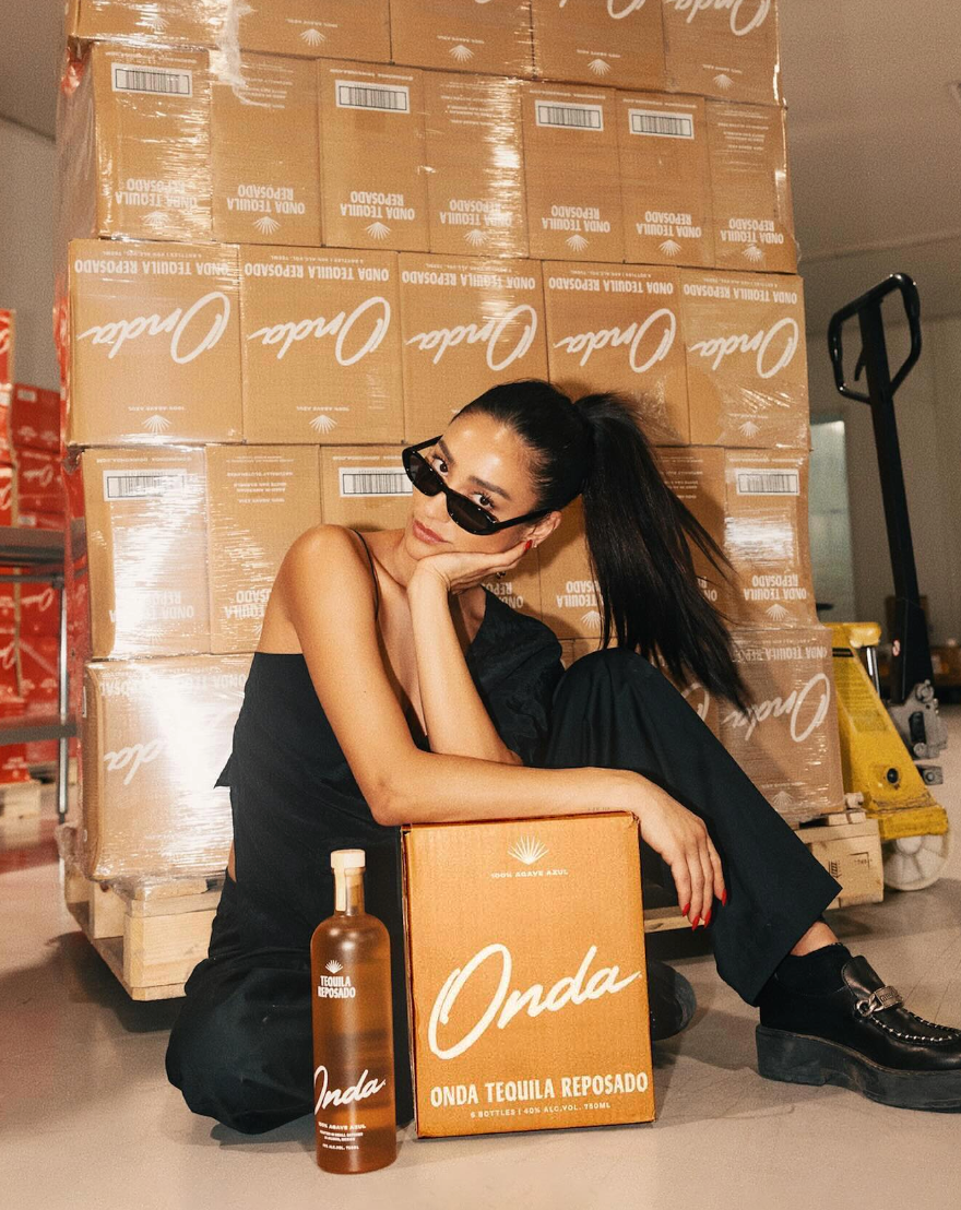 Woman posing with Onda Reposado Tequila box and bottle, surrounded by stacked boxes in a warehouse setting.
