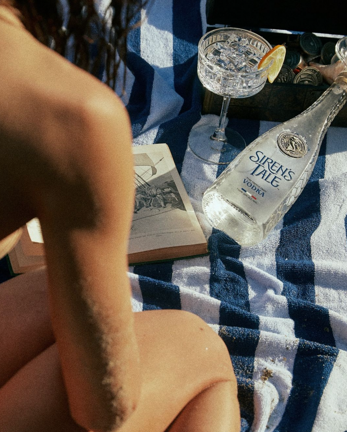 Siren's Tale French vodka bottle displayed on a beach towel with cocktails and books in a sunlit setting.