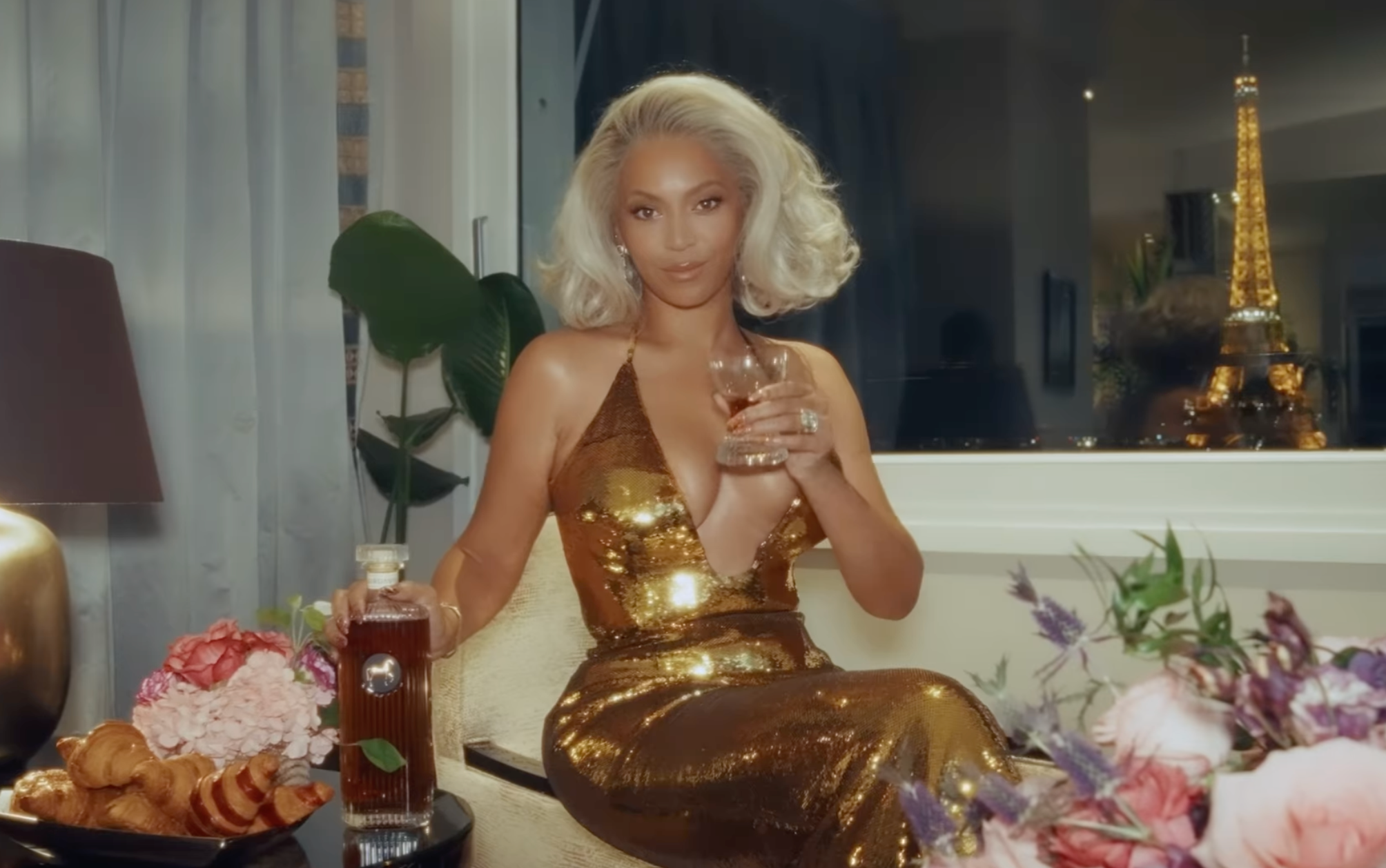 Beyoncé in a glamorous gold dress holding a glass of SirDavis American Whisky, with a Parisian view and elegant decor.