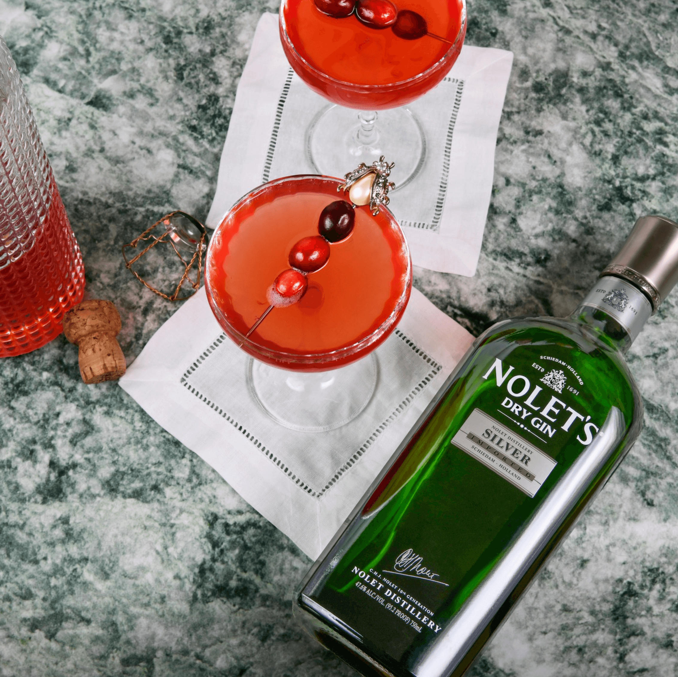 Two cocktails garnished with cranberries beside a bottle of Nolet's Dry Gin Silver on a marble surface.