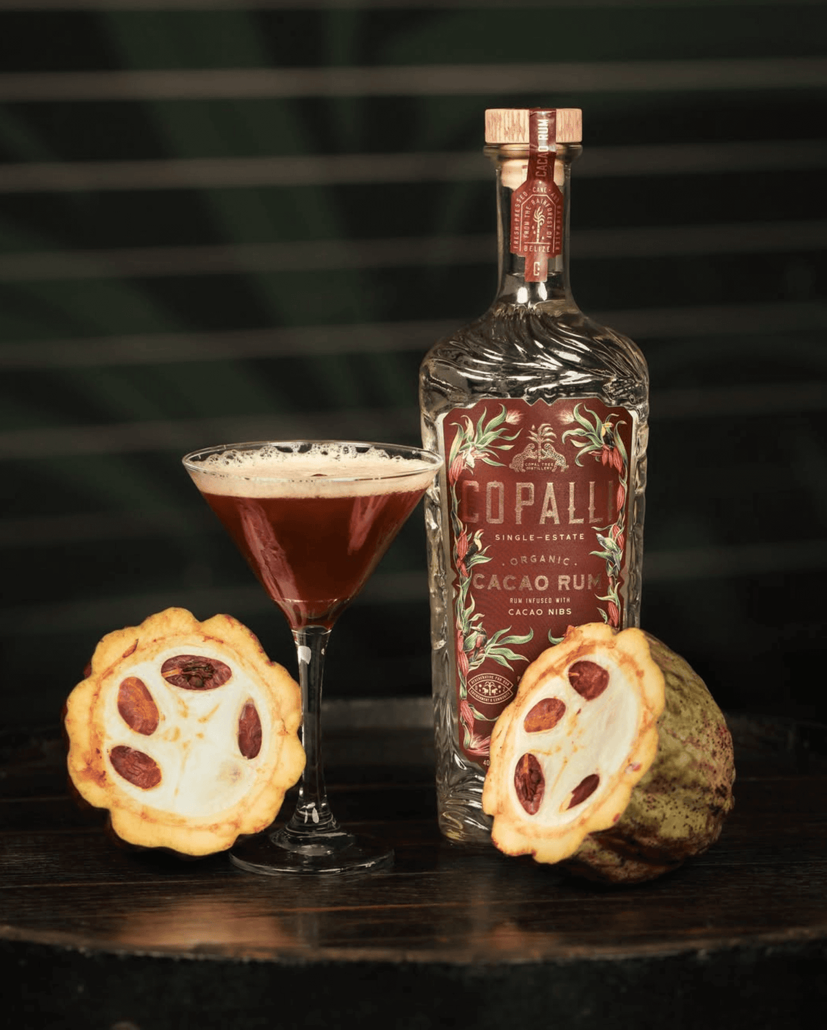 Copalli Cacao Belizean Rum bottle, cocktail in glass, and cacao fruit on wooden table.