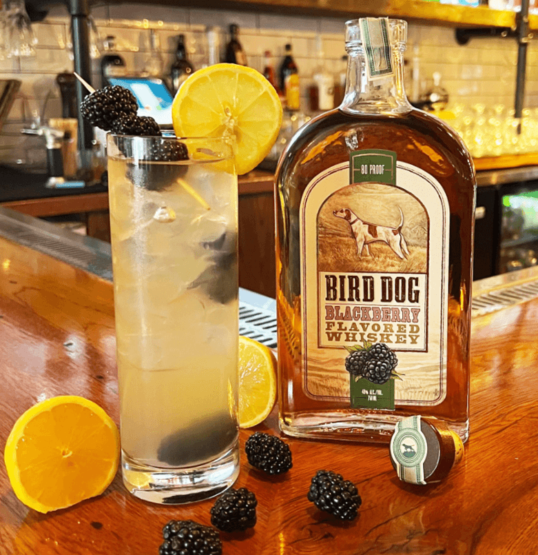 Bird Dog Blackberry Whiskey bottle next to a refreshing cocktail garnished with lemon and blackberries.