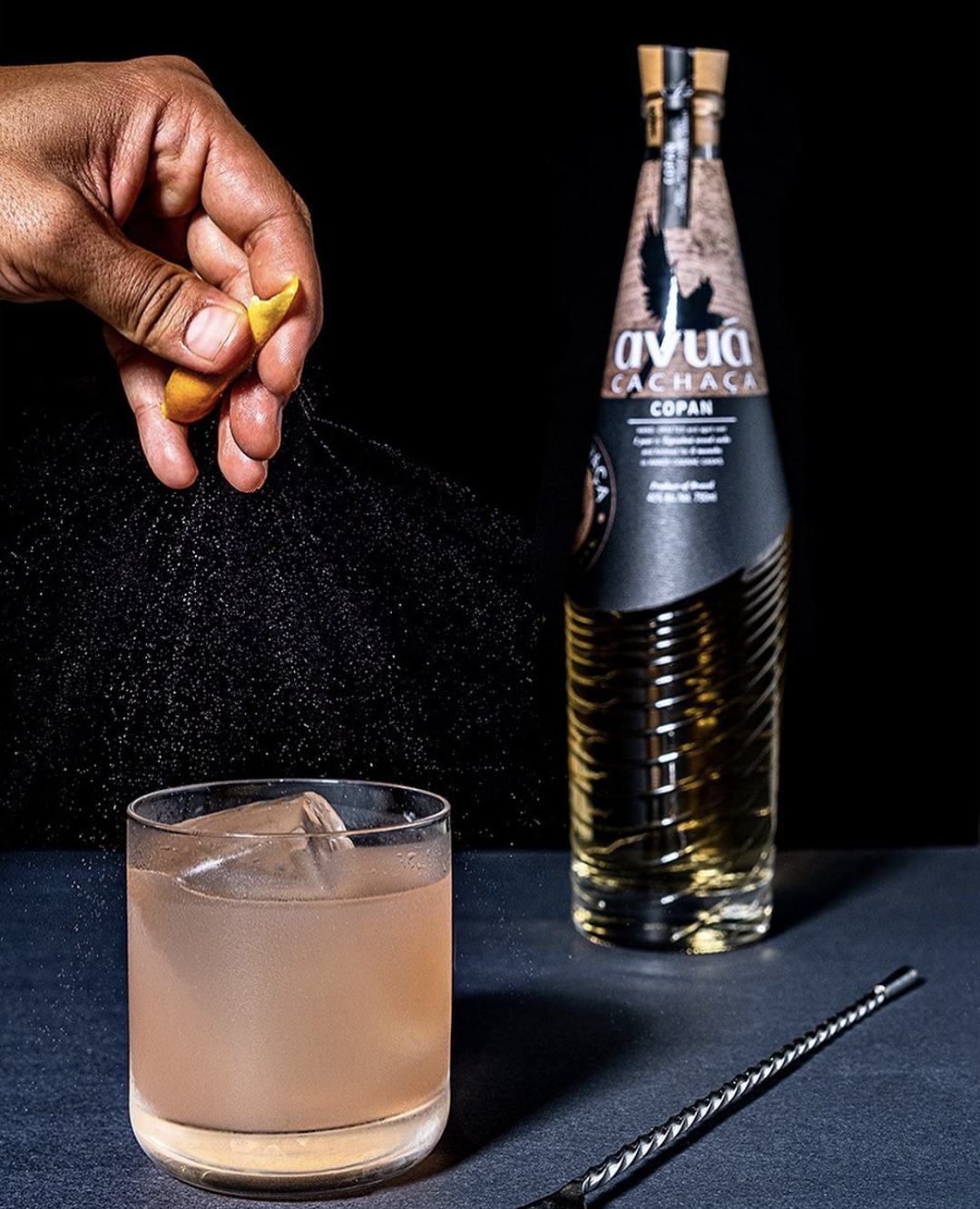 Hand squeezing citrus zest over a cocktail glass beside Avuá Copan Aged Cachaça Limited Edition bottle.