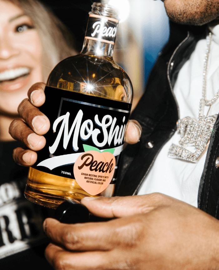 MoShine Peach Moonshine bottle held by a person, featuring a vibrant label and a joyful background.