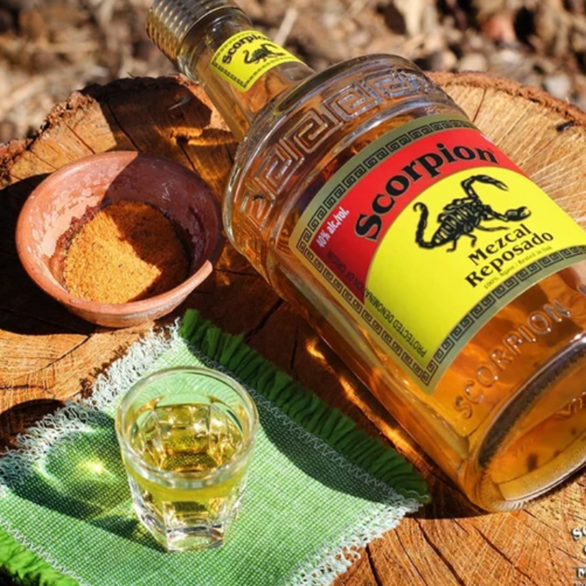 Bottle of Scorpion Mezcal Reposado on a wooden table with a shot glass and chili powder, showcasing rich flavors.