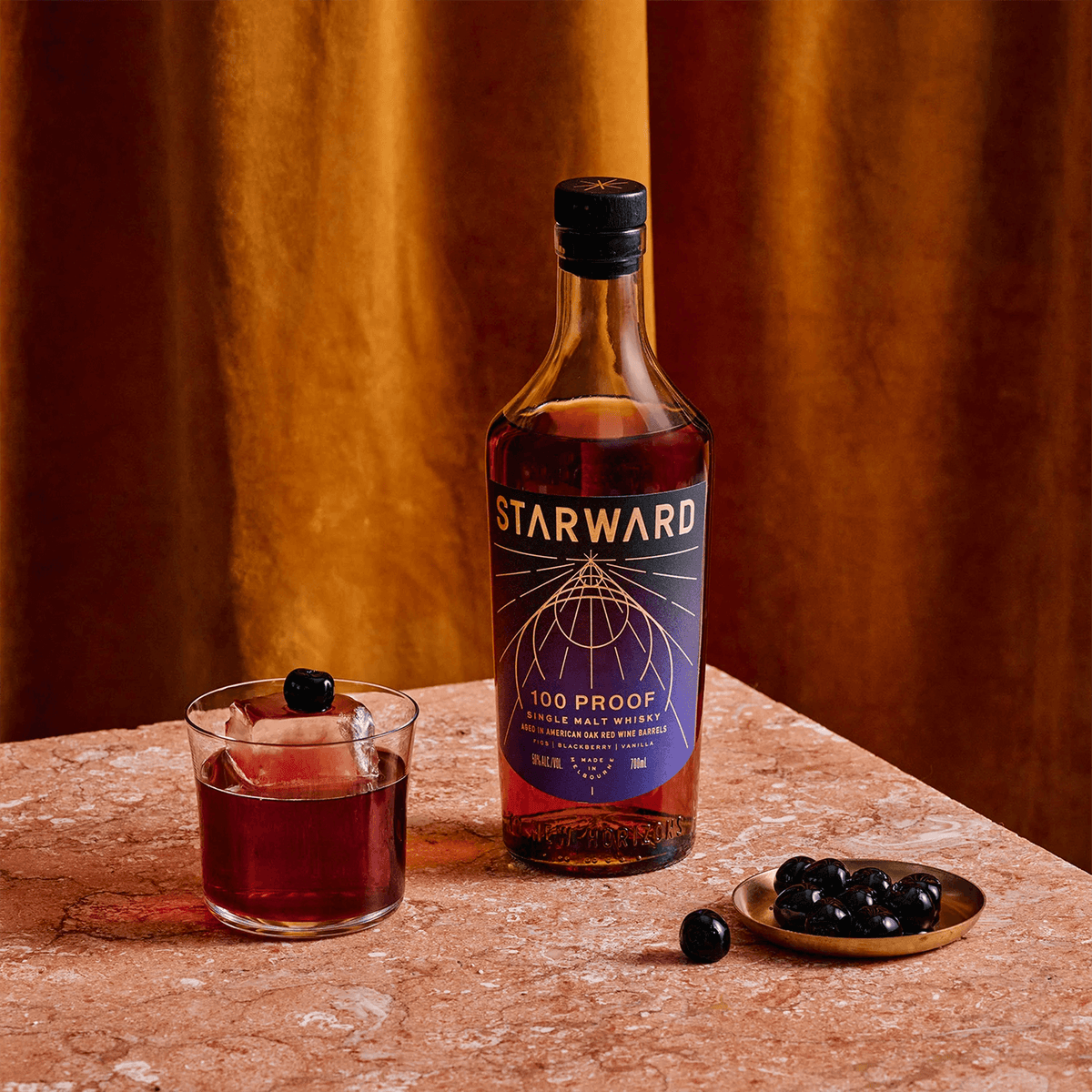 Starward 100 Proof Single Malt Whisky bottle beside a glass of cocktail with berries on a table.