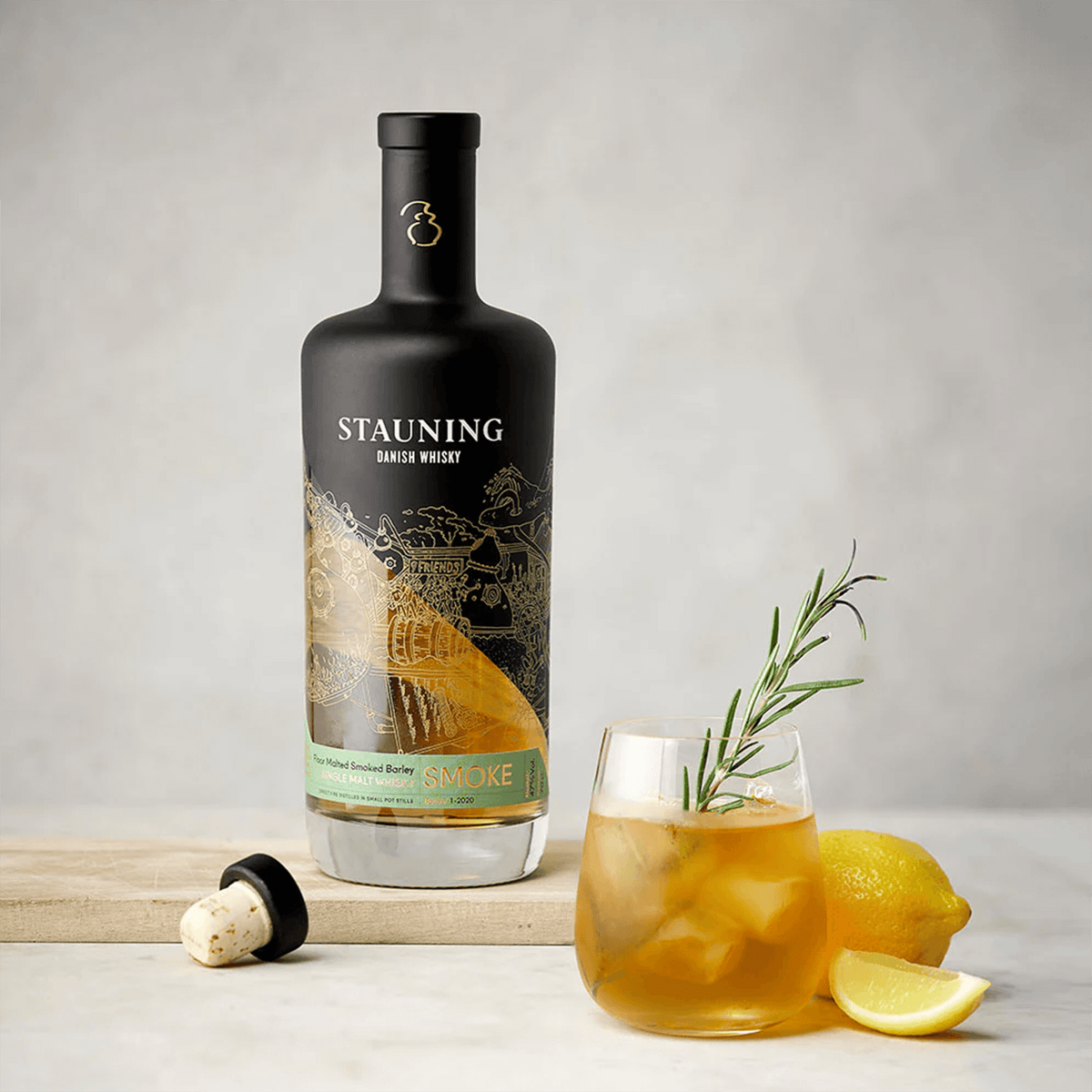 Bottle of Stauning Smoke Single Malt Whisky beside a glass cocktail with lemon and rosemary sprig.
