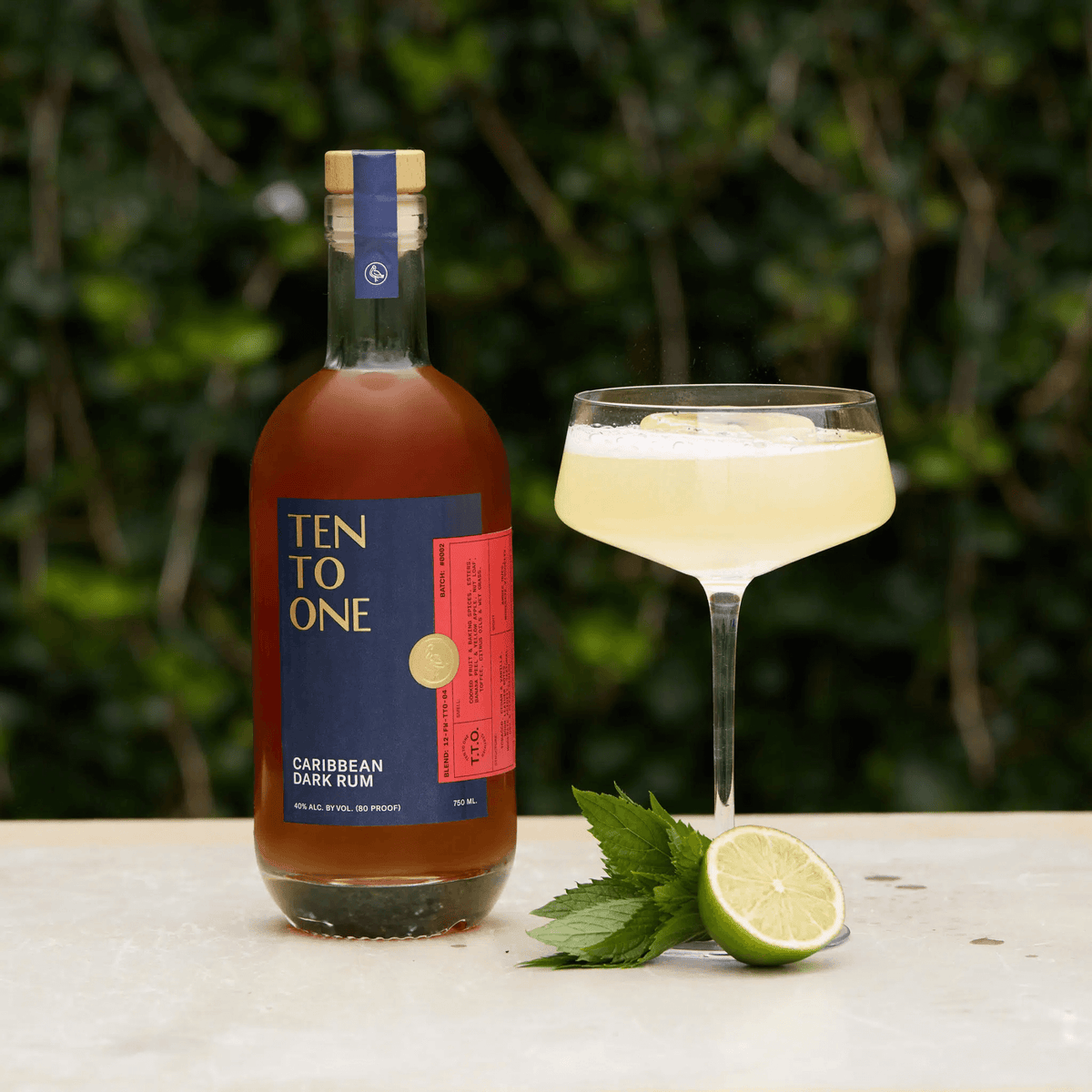 Ten To One Caribbean Dark Rum bottle with cocktail, lime, and mint against a blurred green background.